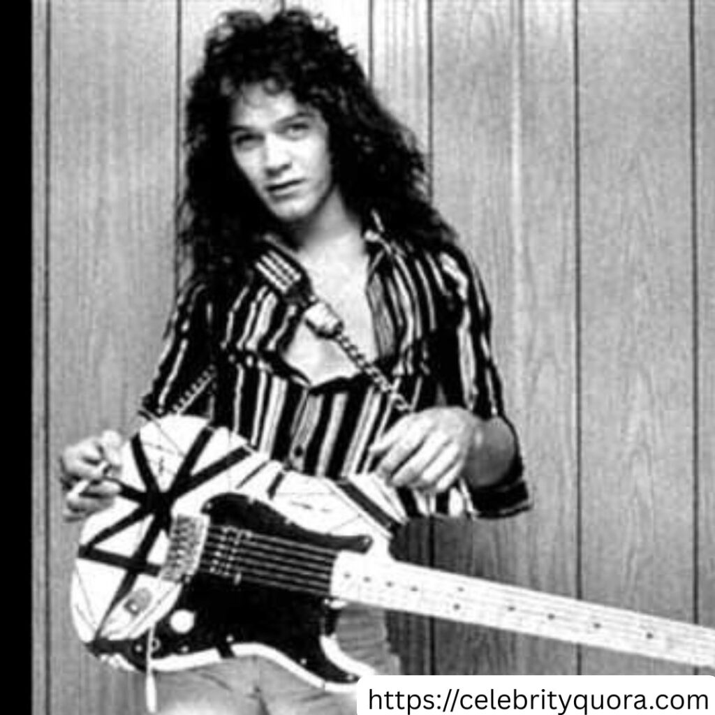 A black and white photo shows Eddie Van Halen holding his striped "Frankenstrat" guitar, looking at the camera. He wears a striped shirt against a wooden plank background.