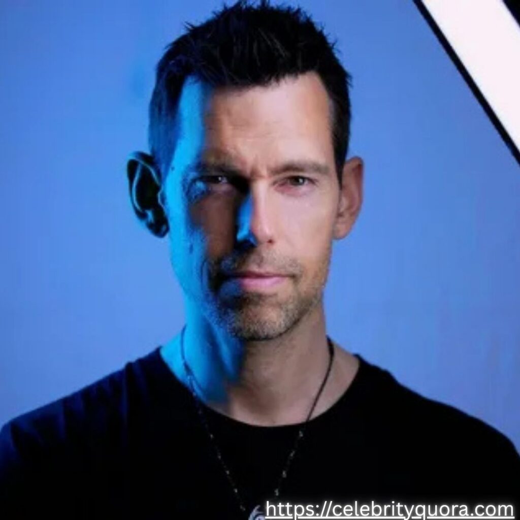 Portrait of Tom Bilyeu with short dark hair and a beard, wearing a black shirt and necklace.
Blue background with a bright light source illuminating him from the right