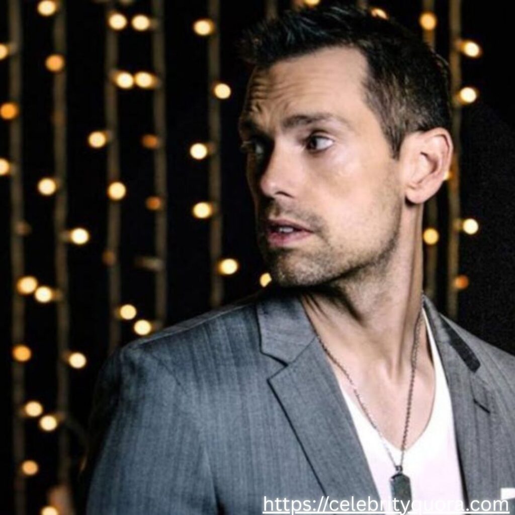 Tom Bilyeu in a grey suit, looking off to the side.
Background of blurred lights.