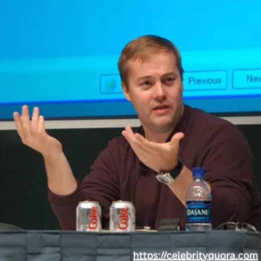 Jason Calacanis gestures with both hands while sitting at a table with two cans of Coke and a bottle of Dasani water.