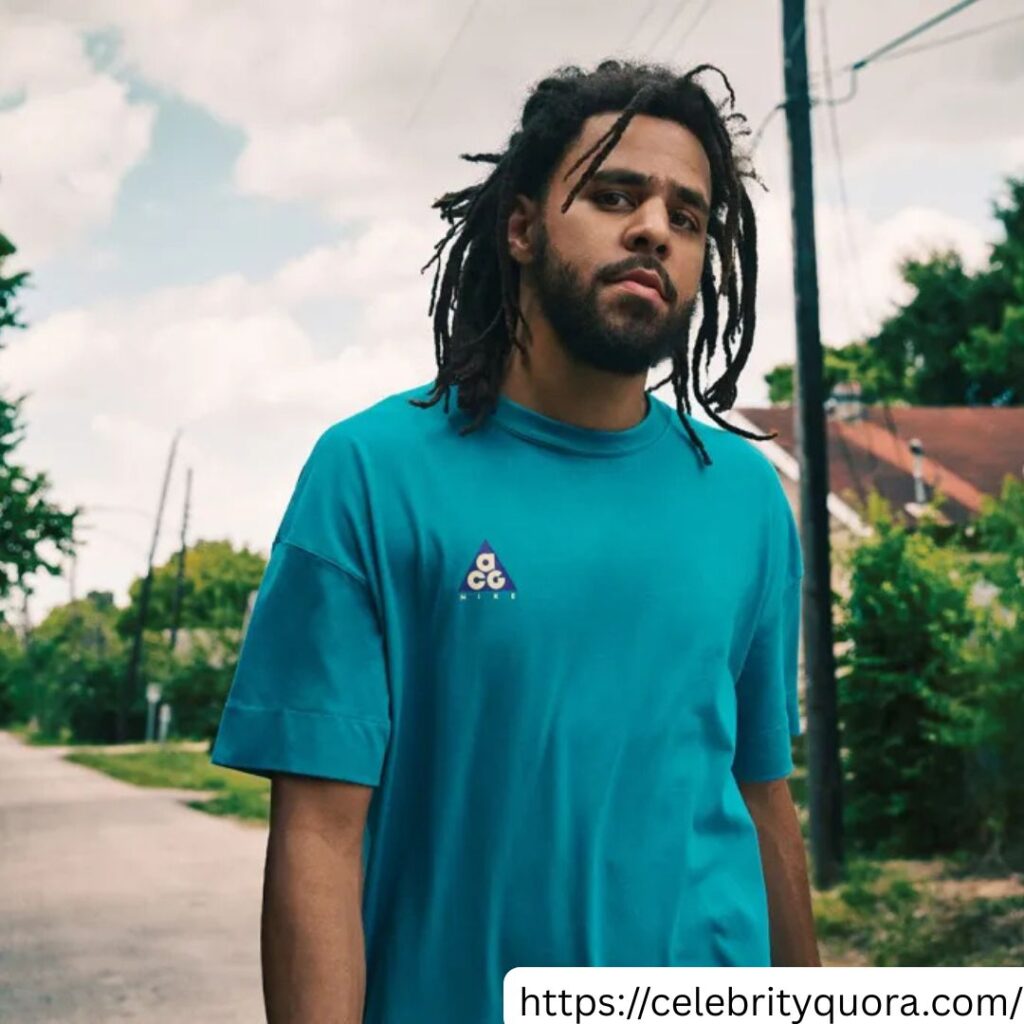 J. Cole is shown on a road, wearing a blue shirt. The Billboard logo is visible in the bottom left corner.

