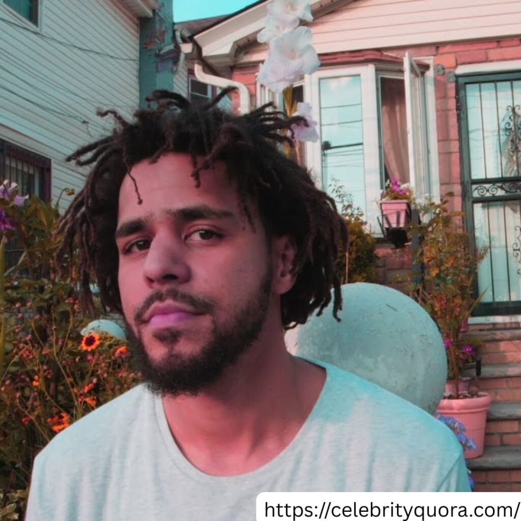  J. Cole, a man who appears to be of African descent, is pictured in front of a brick house with white trim. He has dreadlocks and a beard, and is wearing a light-colored t-shirt.