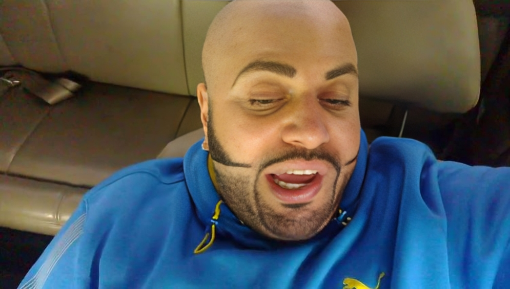  A close-up shot of a bald man with a dark beard and uniquely styled sideburns. He appears to be inside a vehicle, wearing a blue hoodie with a yellow Puma logo.