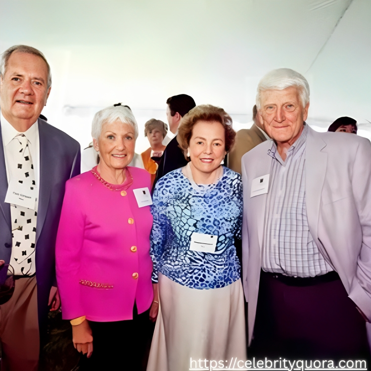 A group of four older adults stand together at an outdoor event. They are dressed in semi-formal attire and appear to be smiling and engaged in conversation.