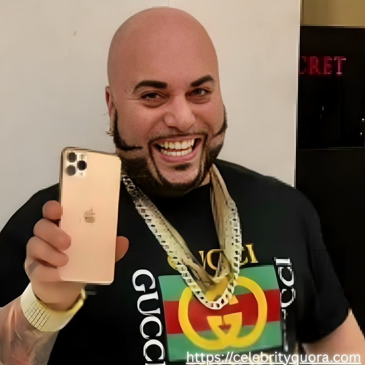 A joyful, bald man with a dark beard and mustache proudly displays a gold smartphone. He is adorned with multiple gold chains, a watch, and a bracelet, wearing a Gucci t-shirt and patterned jeans in what seems to be a shopping mall.