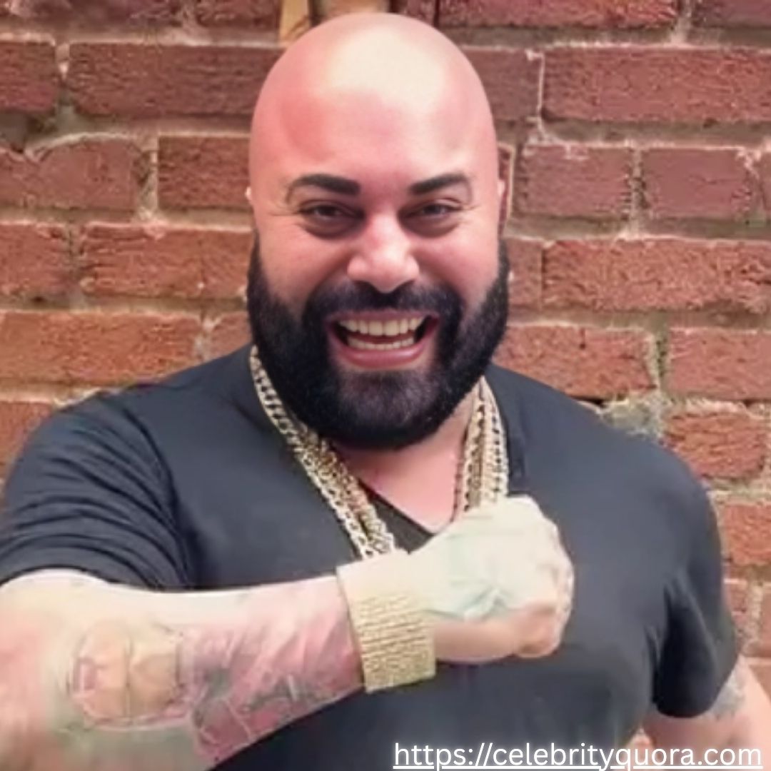 A bald man with a dark beard laughs heartily while wearing multiple gold chains and a gold watch. He is wearing a dark t-shirt and has tattoos on his left forearm and hand. He is standing against a brick wall.