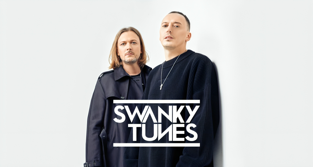 Please write an alt text for this image   Two light-skinned men stand against a stark white background. The man on the left has shoulder-length blonde hair and a short beard; he's wearing a dark trench coat. The man on the right has short, light brown hair and is wearing a black sweater and a silver cross necklace. The words "SWANKY TUNES" are prominently displayed in a stylized black and white font in the lower center of the image.