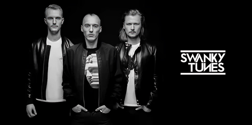 A black and white shot features three light-skinned men against a solid black background. The man on the left has short, light hair and is wearing a light-colored shirt under a dark leather jacket. The man in the center is bald and wears a dark jacket over a patterned shirt. The man on the right has shoulder-length light hair and is wearing a light-colored shirt under a dark leather jacket. To the right, the words "SWANKY TUNES" are displayed in a white, stylized font.