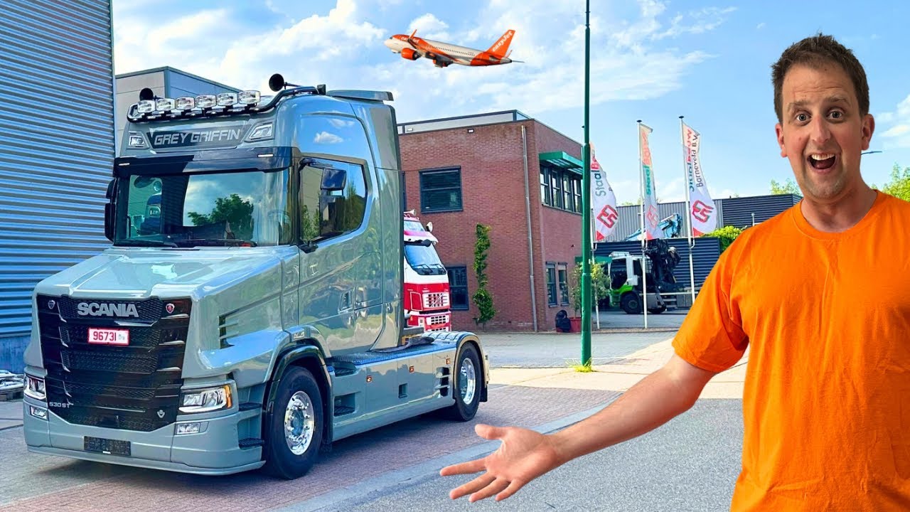A man in an orange t-shirt stands excitedly in front of a sleek, silver Scania truck parked on an asphalt lot. In the background, buildings, flags, and a bright blue sky with a flying airplane complete the scene.