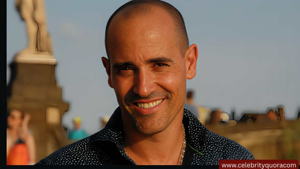 A smiling man with a shaved head and a patterned blue shirt looks directly at the camera. In the blurred background, a statue and other people suggest an outdoor setting.
