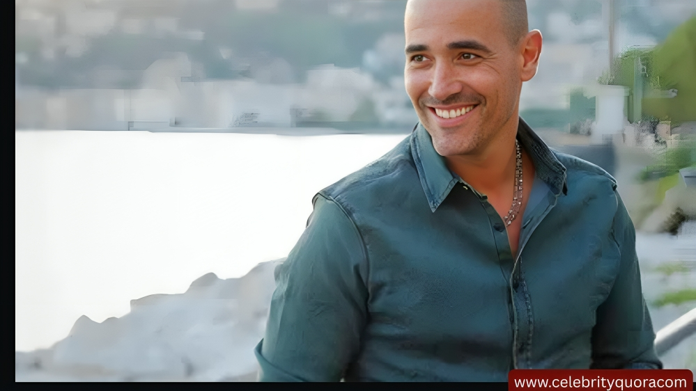 David Rocco smiles with a city and ocean view behind him. He is wearing a casual blue shirt and bracelets.