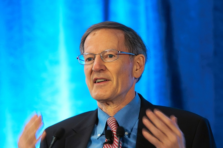 A medium shot captures George Gilder, a distinguished older white man with short brown hair and glasses, speaking with his hands raised in a gesturing motion. He is wearing a dark suit jacket over a light blue collared shirt and a diagonally striped red and white tie. Two microphones are clipped to his tie. Behind him is a blurred backdrop of vibrant blue curtains.