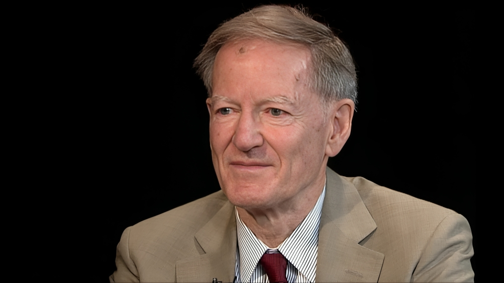 A close-up shot features George Gilder, an older white man with short, light brown hair. He is wearing a light brown suit jacket over a white collared shirt with thin blue stripes and a dark red tie. He is looking slightly to his left with a subtle smile. The background is a solid, dark black.