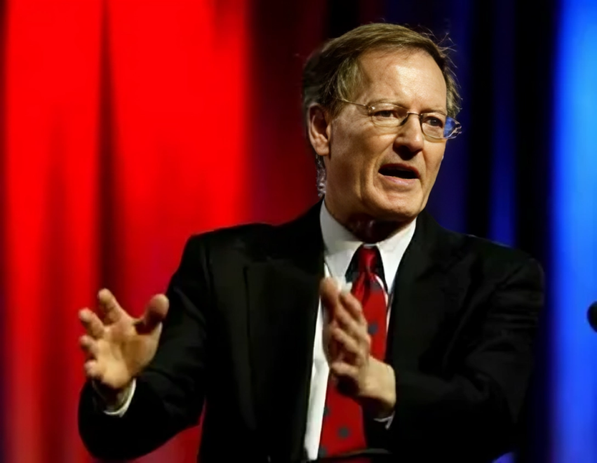 Please write an ALT text for this image   A medium shot shows George Gilder, an older man who appears to be white, with short brown hair and glasses, speaking with animated hand gestures. He is dressed in a dark suit jacket, a white collared shirt, and a red tie with white patterns. The background features blurred red and blue curtains. A microphone is visible to the right of the frame.