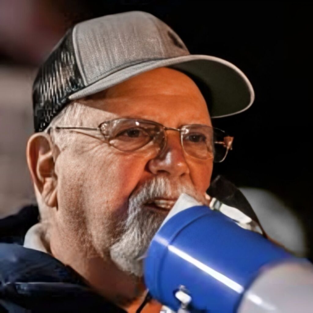 A close-up shot of Harold Daggett,  with a white beard, speaking into a megaphone while wearing a baseball cap.