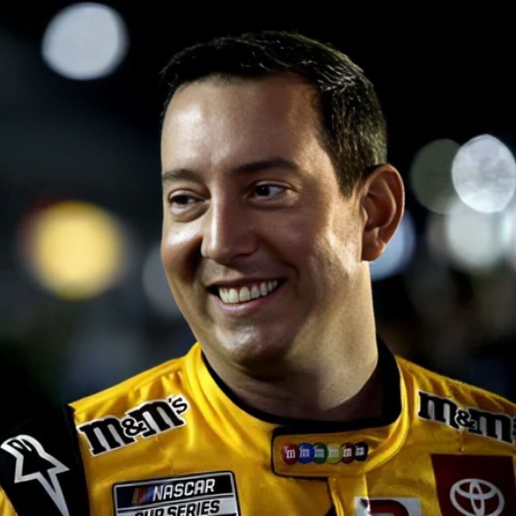 A close-up shot of Kyle Busch smiling and wearing sunglasses, a black baseball cap, and a blue and green race suit.