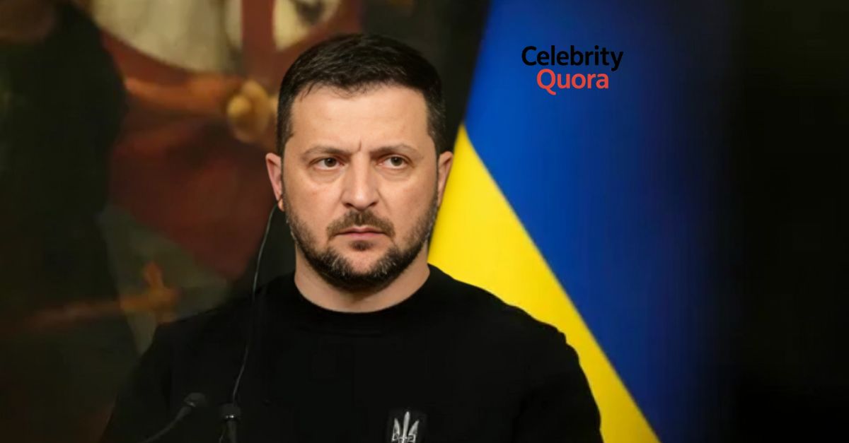 Ukrainian President Volodymyr Zelenskyy, with a beard and dressed in a dark top, looks intently forward, with a Ukrainian flag visible in the background.