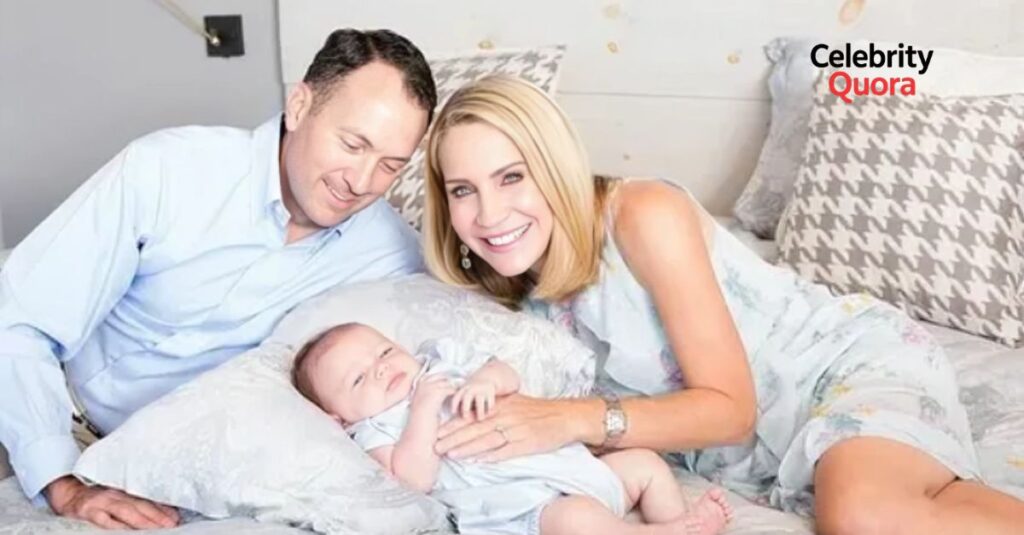 Andrea Canning smiling and lying on a bed next to her husband and a baby. her husband is with short brown hair, is looking down at the baby with a smile. Andrea is wearing a light blue dress with a white pattern. The baby is lying on a pillow in a light blue outfit.