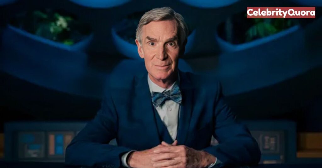 A medium shot of Bill Nye sitting at a futuristic-looking desk with his hands clasped in front of him. He is wearing a dark blue suit jacket, a white shirt, and a patterned blue bowtie. The background is dark and features circular, futuristic-looking panels with a hint of green foliage inside some of them.
