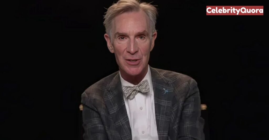 A head-and-shoulders shot of Bill Nye, the science communicator, smiling at the camera. He is wearing a gray plaid suit jacket, a white shirt, and a light-colored bowtie with a subtle pattern.