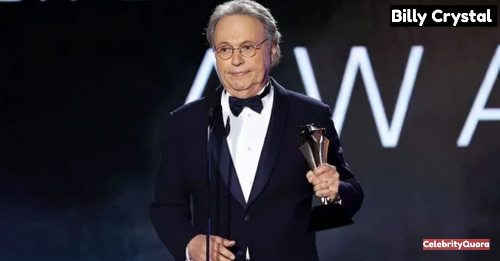 Billy Crystal, wearing a navy blue tuxedo with a black bow tie and glasses, stands on a stage holding a gold award. He's looking slightly to his left with a thoughtful expression. The background is dark with blurry, large white letters that appear to spell "AWARDS."