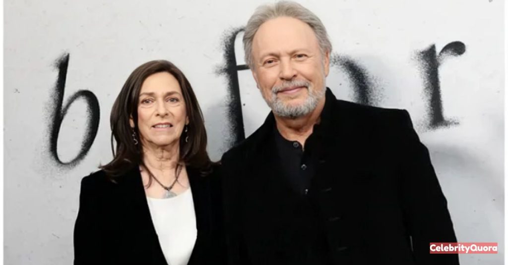A photo of Billy Crystal and his wife, Janice Goldfinger, smiling at the camera. Billy is wearing a black collared jacket, and Janice is wearing a similar black jacket over a white top. They are standing in front of a white wall.