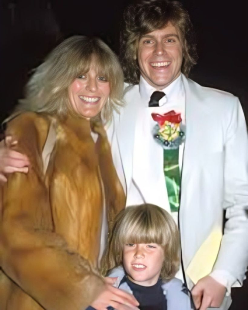 A smiling Jeff Conaway in a white tuxedo with a red rose and green foliage corsage, standing with his arm around his smiling blonde wife, who is wearing a tan fur coat. Their son stands in front of them, also smiling, wearing a blue and white jacket.