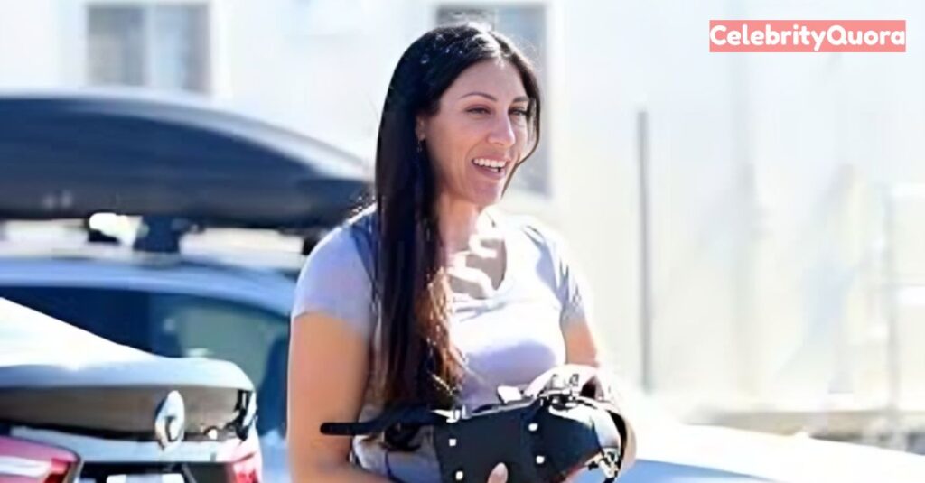 
Kristin Grannis smiles, looking to her left, while holding a black bag in front of her. She is wearing a light-colored t-shirt, and her long, dark hair is down.