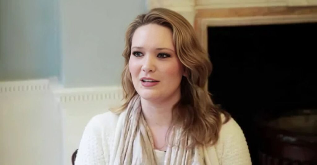 A smiling Sarah J. Maas, the author of fantasy novels, is seen in a head-and-shoulders portrait. She has long, wavy blonde hair and is wearing a light-colored sweater and scarf.