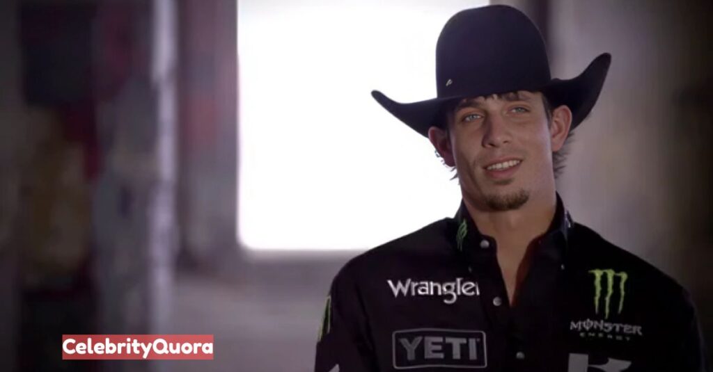 A portrait of J.B. Mauney, a bull rider, smiling slightly and wearing a black cowboy hat and a black long-sleeved shirt with sponsor logos.