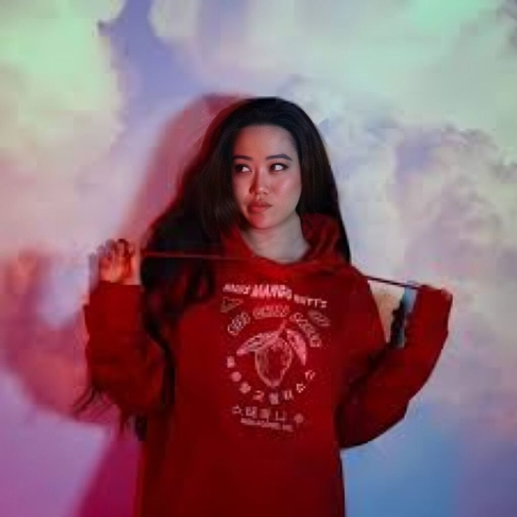 Stephanie Soo posing in a red hoodie with a graphic design of a mango on it. She is holding the drawstrings of the hoodie and looking to the side. The background is a soft, multicolored cloud-like pattern.