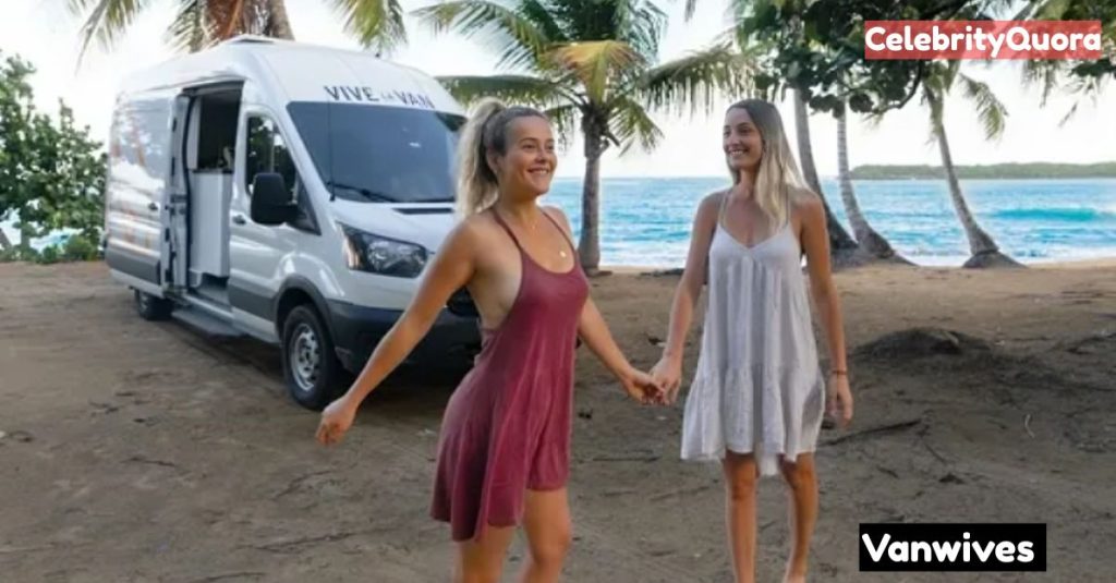 Jazmyn Canning & Crystal Drinkwalter holding hands and walking toward the camera on a sandy beach. A white camper van and palm trees are behind them, with the ocean in the distance.