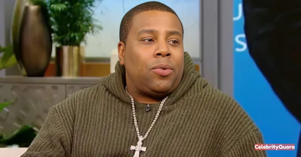 Kenan Thompson in a casual, thoughtful pose, wearing a green hooded sweatshirt and a large silver cross necklace.