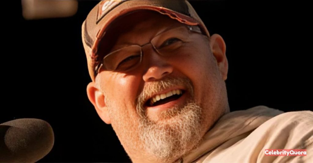 A close-up shot of comedian Larry the Cable Guy smiling and laughing, wearing a light-colored jacket and a camouflage-patterned baseball cap.