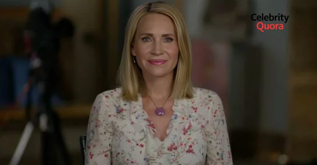 A medium shot of Andrea Canning, a blonde woman with blue eyes, looking directly at the camera with a slight smile. She is wearing a white floral blouse and a purple pendant necklace. The background is a slightly blurred, indoor setting with a camera tripod visible on the left.