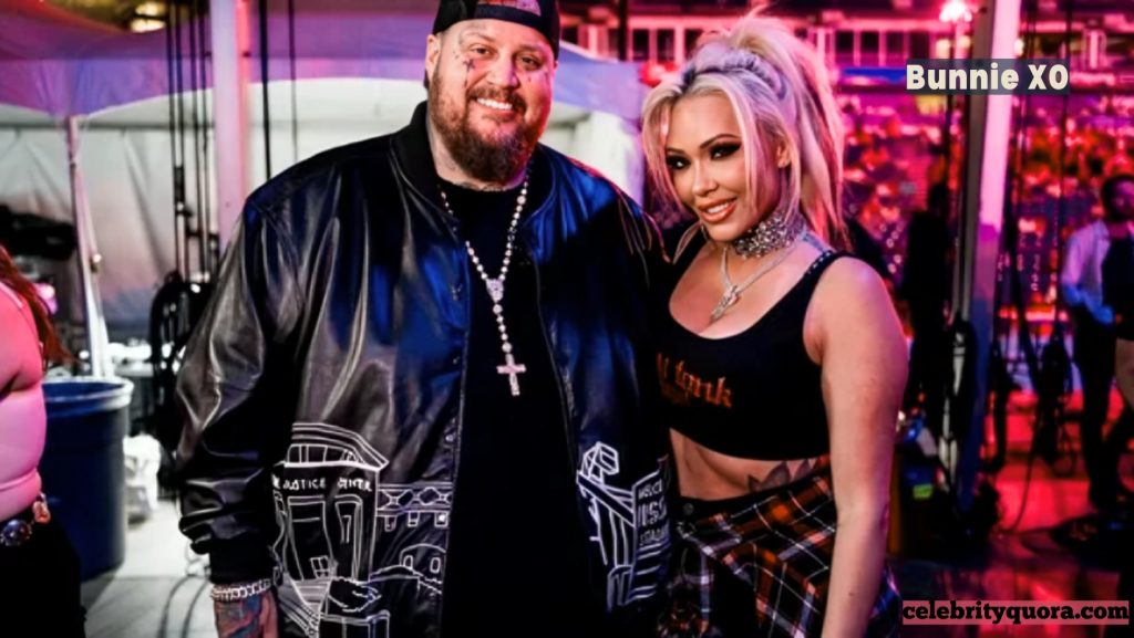 Bunnie XO posing with her husband, Jelly Roll, backstage. She is wearing a black crop top and a plaid skirt, while he is dressed in a black leather jacket and a backwards baseball cap. They are both smiling for the camera.