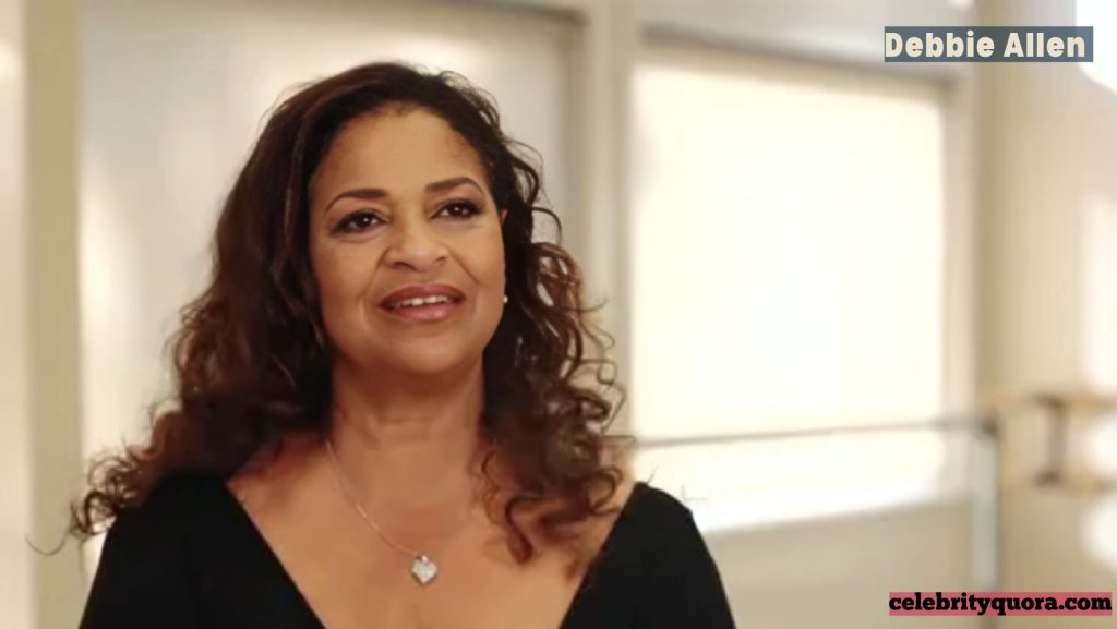 A recent photo of Debbie Allen, with long, wavy brown hair, wearing a black top and a silver heart pendant necklace, smiling indoors.