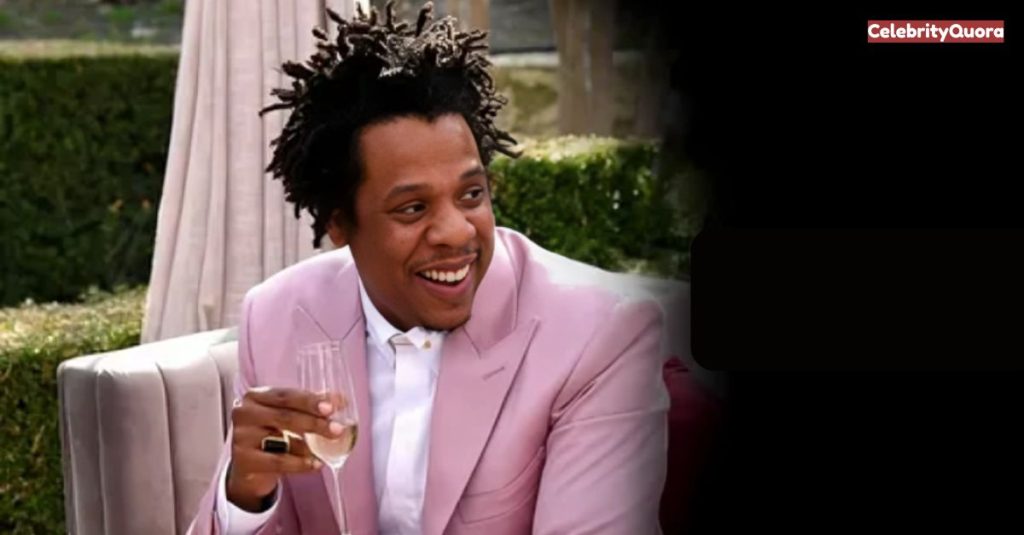Jay-Z smiling, holding a glass of champagne while wearing a pink suit.