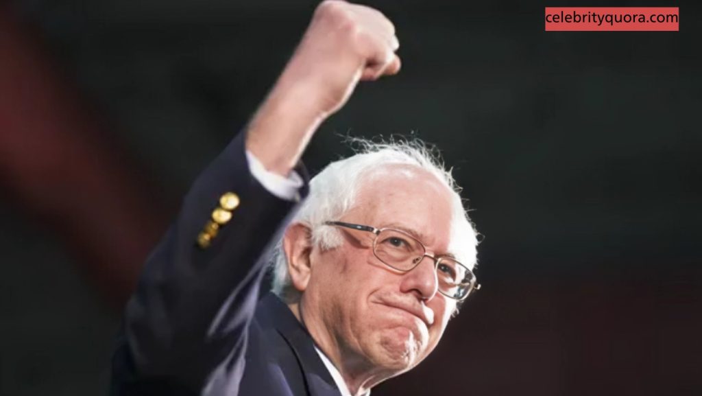 A close-up of a smiling Bernie Sanders, wearing a dark suit and glasses, with his right arm raised in a triumphant fist. The background is a dark, out-of-focus red.