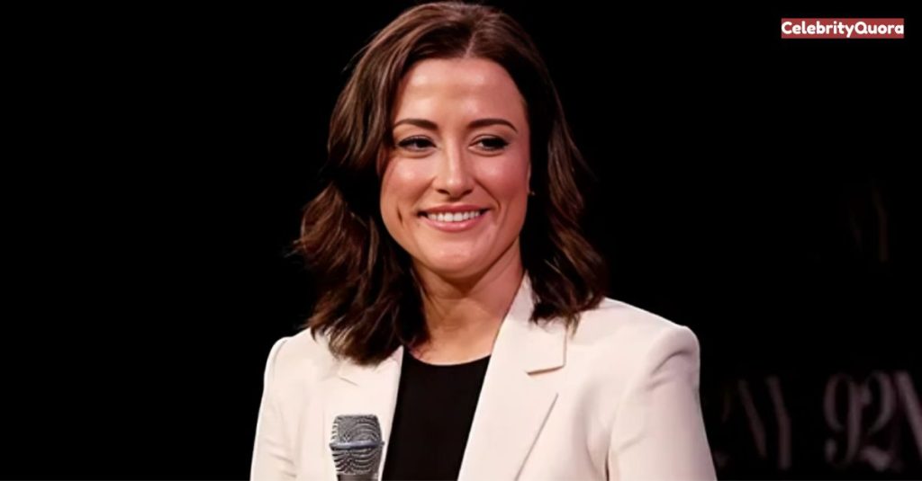 Cassidy Hutchinson smiles during an interview on a television show. She is wearing a dark, sparkling top and a pair of earrings. In the background, there is a blurry city skyline at night.