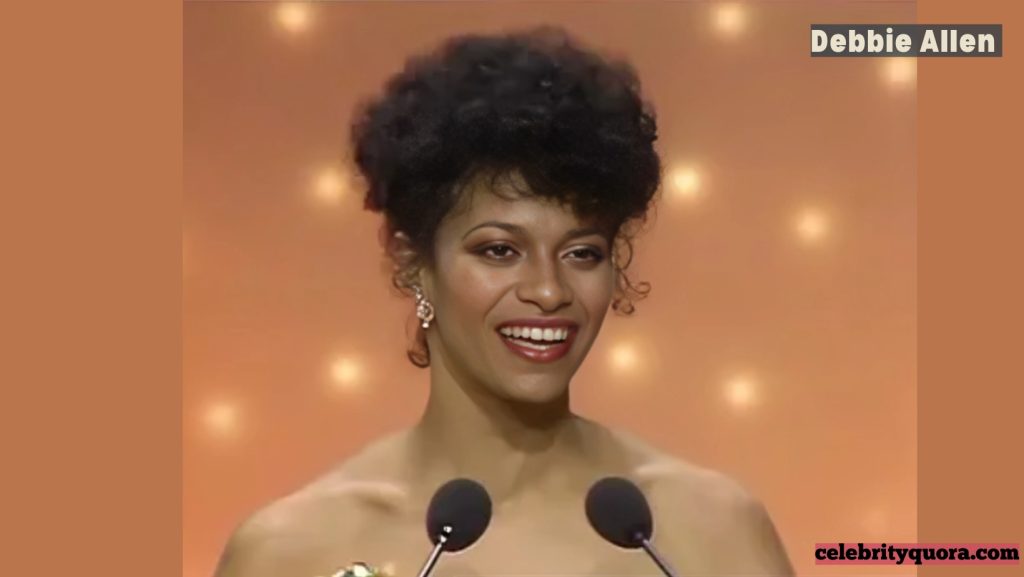 Headshot of actress and choreographer Debbie Allen in her younger years, standing on a stage with a golden background and two microphones in front of her.