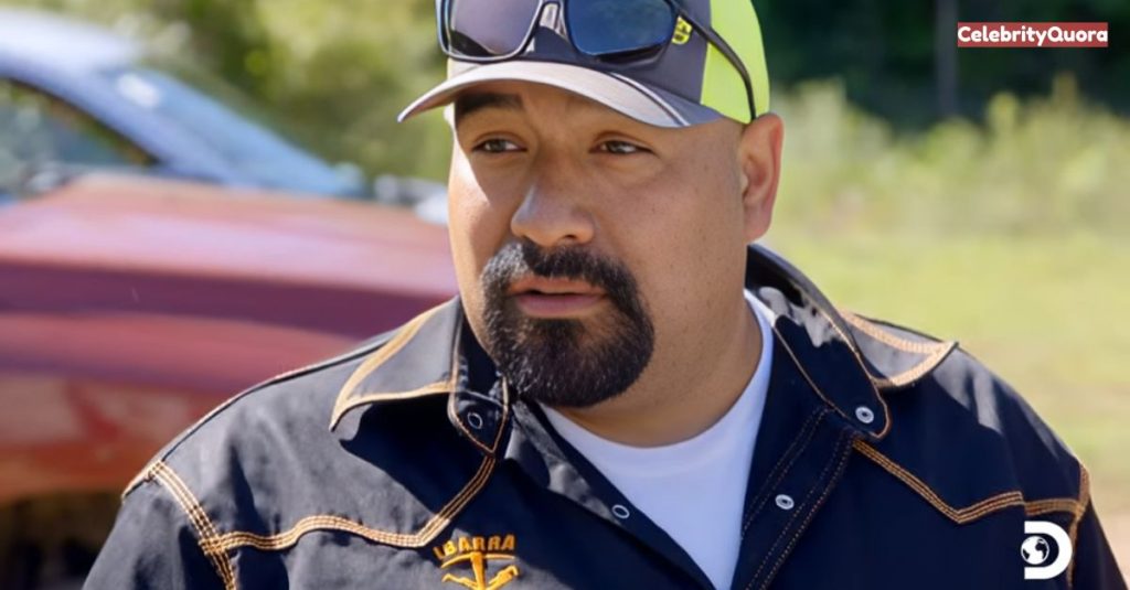 Juan Ibarra is shown outdoors, wearing a black work jacket with an "Ibarra" logo and a white shirt underneath. He's also wearing a cap and sunglasses on top of his head. He has a goatee and is looking forward with a serious expression. A vehicle is partially visible in the blurry background.
