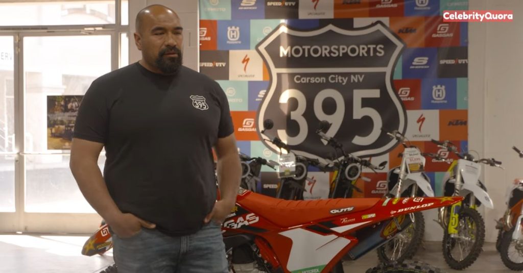 Juan Ibarra, stands in what appears to be a motorsports showroom. He is wearing a black t-shirt with a logo and jeans, with his hands in his pockets. He has a beard and is looking slightly to his left. In the background, there are several motorcycles and a large sign for "395 Motorsports Carson City NV.