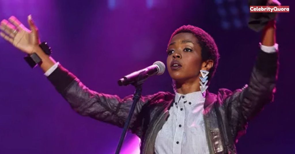 Lauryn Hill on stage with her hands raised in the air, a microphone in her right hand. She is wearing a black jacket and a black and white striped shirt, and her hair is short. The background is a mix of purple and pink stage lights.