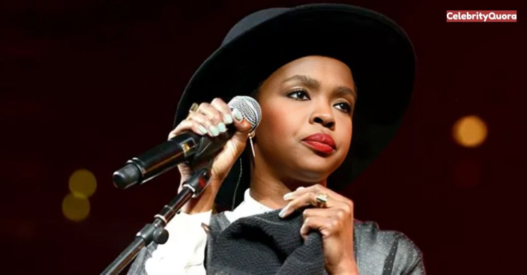 A close-up shot of Lauryn Hill on stage, wearing a black hat and a white shirt with a dark jacket. She is holding a microphone with her left hand and a dark cloth with her right, looking slightly to the side with a serious expression.