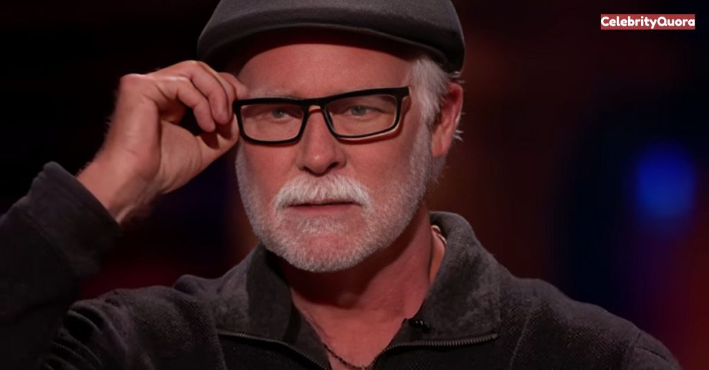 A close-up portrait of Mark Singer Gorilla Glue with a gray goatee and mustache. He is wearing a black hat and a dark gray sweater and is in the process of putting on or taking off his glasses. He has a serious expression, and his blue eyes are visible.

