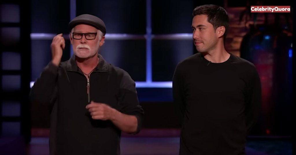  A scene from "Shark Tank," showing Mark Singer of Gorilla Glue and his business partner on stage during their presentation. Mark, with his signature gray goatee and black hat, makes a point with a hand gesture. His partner stands quietly by his side.